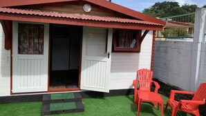 A small, single-story building with a red roof and white walls, featuring a door and windows, and two red plastic chairs outside. - Bungalow for 2 persons (Les Trois-Îlets)