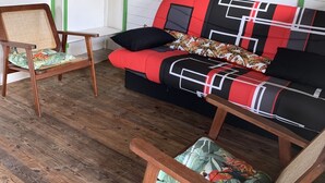 A room with a wooden floor, a red and black sofa with patterned cushions, and two wooden chairs with woven seats. - Bungalow for 2 persons (Les Trois-Îlets)