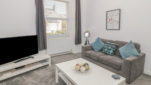 A living room with a flat-screen TV, a white coffee table with decorative items, a grey sofa with patterned cushions, and a framed artwork on the wall. - Apartment 2 (Llandudno)