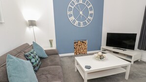 A modern living room with a brown sofa, teal cushions, a white coffee table, a large wall clock, a TV on a white stand, and a fireplace with logs. - Apartment 2 (Llandudno)
