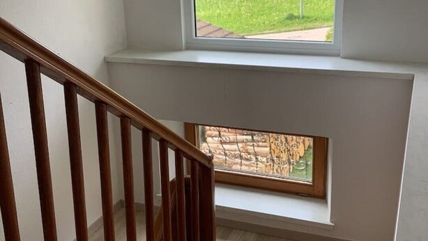 Eine Treppe mit hölzernen Geländern führt zu einer Zwischenebene mit einem Fenster mit Blick auf ein grünes Feld.