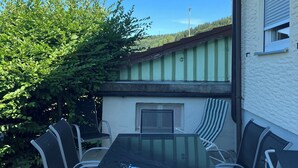 Eine Außenpatio mit einem Tisch aus Glasplatten und Metallstühlen, umgeben von Grünpflanzen und einem Gebäude mit einer Gründachfläche.