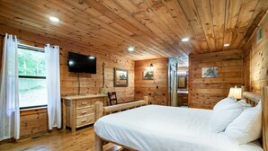A wooden cabin room with a bed, a desk, a chair, a TV, and a window with curtains. - Private Cabin Retreat – Relax in the Hot Tub & Cozy Up by Fire! (Pigeon Forge)
