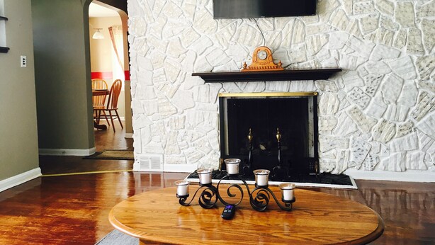A living room with a stone wall, a mounted TV, a fireplace with a mantel, and a round wooden table with candles and a remote control. - Cozy & Quiet beautiful 4 bedrooms , close to downtown, Cleve clinic  (Parma)