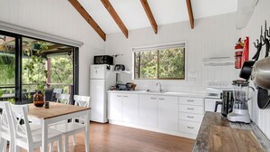 A modern kitchen with white cabinetry, a wooden dining table, and a view of greenery outside. - Refreshing Rainforest Retreat - Kangaroo Valley (Kangaroo Valley)