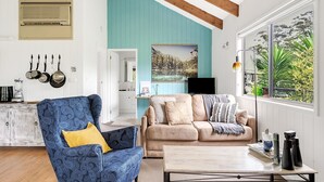 A living room with a blue armchair, beige sofa, and a coffee table. There is a painting on the wall, a window with a view of greenery, and a kitchen area with hanging pots. - Refreshing Rainforest Retreat - Kangaroo Valley (Kangaroo Valley)