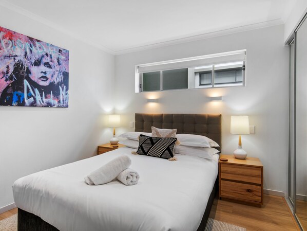 A modern bedroom with a large bed, two bedside tables with lamps, a headboard, a window with blinds, and a wall-mounted painting.