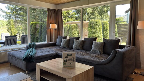 A modern living room with a sectional sofa, a coffee table, and large windows offering a view of a garden. - Geräumige Wohnung in Altenau mit Terrasse (Altenau)