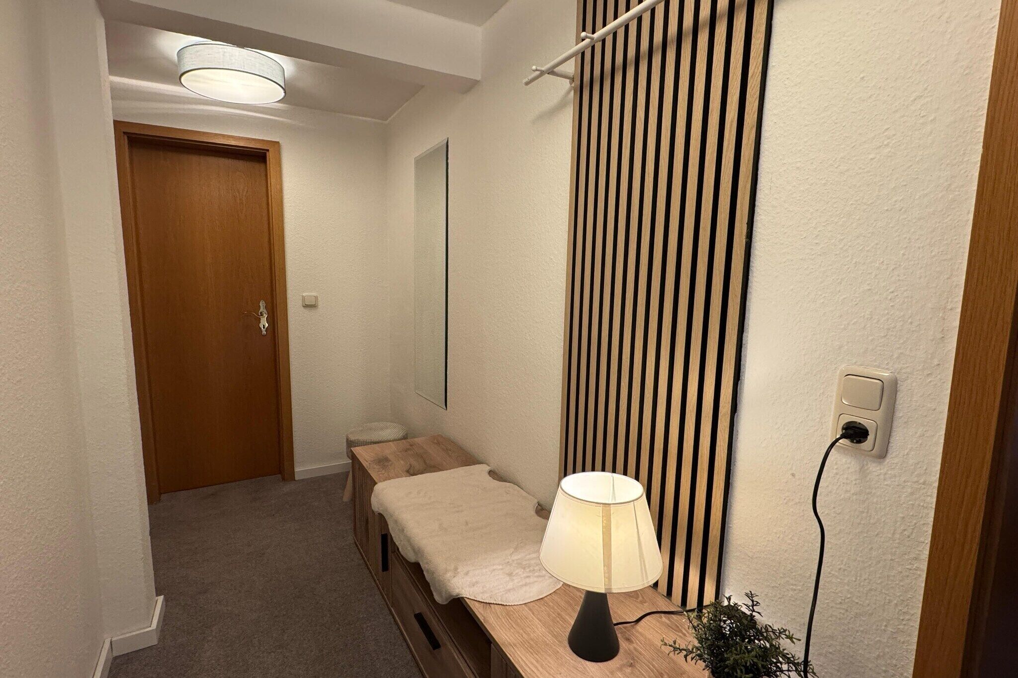 A small, cozy room with a wooden door, a bedside table with a lamp, a small stool, and a vertical slat wall design.