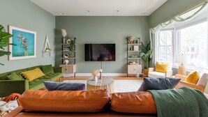 Un salon avec un canapé vert, un pouf en cuir orange et un téléviseur à écran plat. On y trouve des étagères ornées d’objets décoratifs et une fenêtre avec des rideaux.