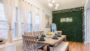 Un coin repas avec une table en bois dressée pour quatre personnes, entourée de chaises, d’un banc et d’un panneau néon mural sur lequel on peut lire « Boho House ».