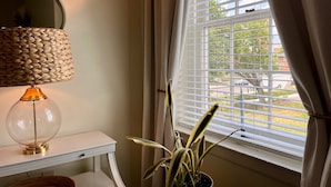 Una habitación con una lámpara sobre una mesa blanca, una planta en maceta y una ventana con persianas.
