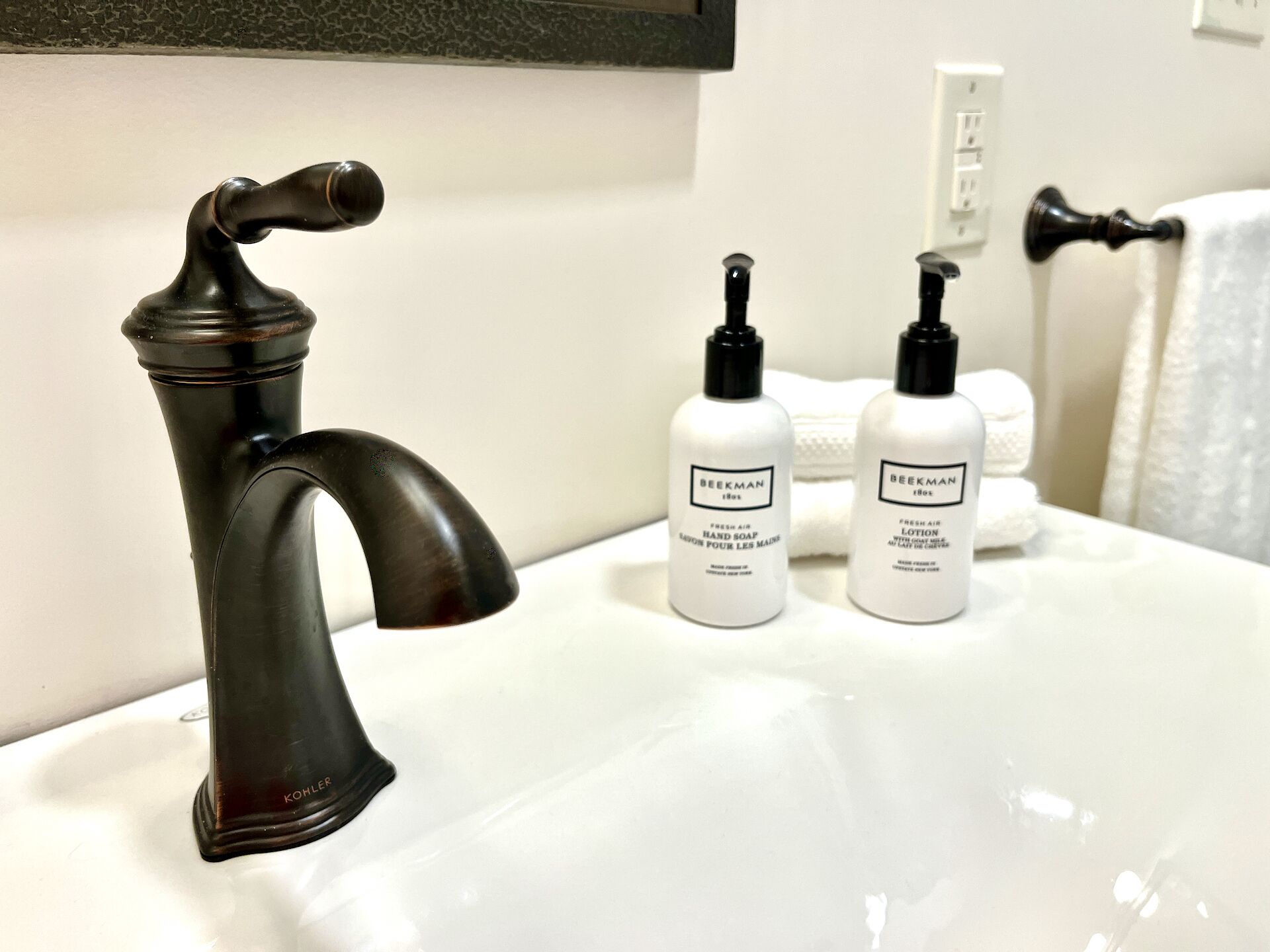 A bathroom with a black faucet, two bottles of Beekman hand soap and lotion, and a towel rack with a white towel.