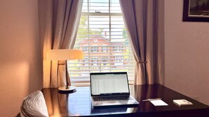Quarto de hotel com escrivaninha, laptop, luminária e vista para edifícios pela janela.