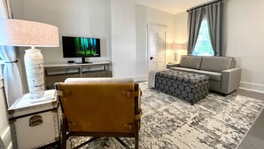 Ein Hotelzimmer mit Fernseher, einer Couch, einem Sessel, einem gemusterten Teppich und einem Fenster mit Vorhängen.