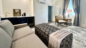 Ein modernes Hotelzimmer mit einer Couch, einem gemusterten Hocker, einem kleinen Tisch mit Stuhl, einer Küchenzeile mit Spüle und Kaffeemaschine sowie einem großen Fenster mit Vorhängen.