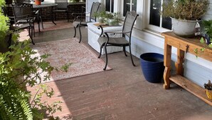 A covered porch with a brick floor, wooden furniture, and potted plants.
