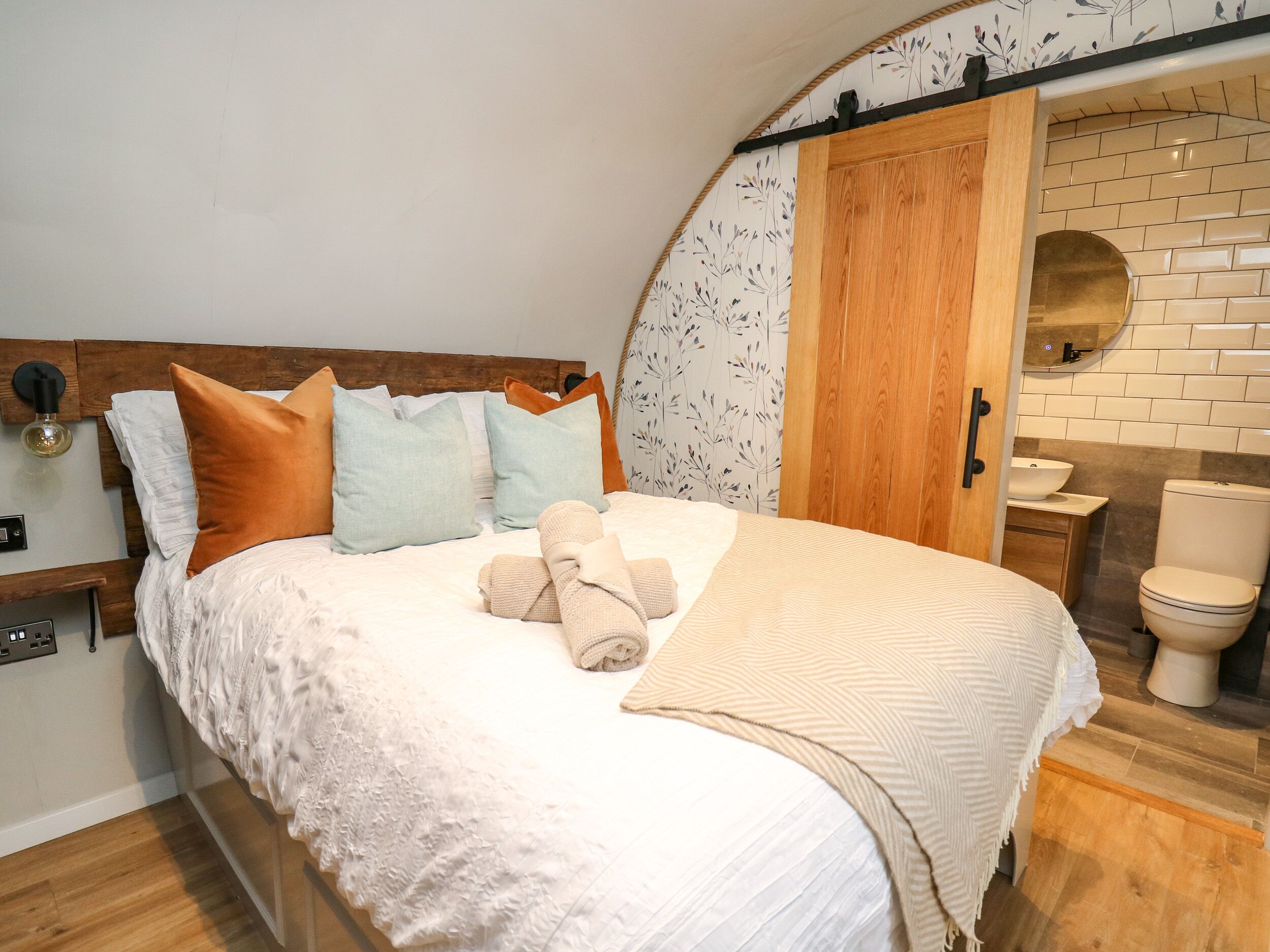 Un dormitorio con cabecera de madera, una cama con ropa de cama blanca y varios almohadones, una mesita de noche con lámpara, y vistas a un baño con lavamanos e inodoro.