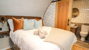 Un dormitorio con cabecera de madera, una cama con ropa de cama blanca y varios almohadones, una mesita de noche con lámpara, y vistas a un baño con lavamanos e inodoro.