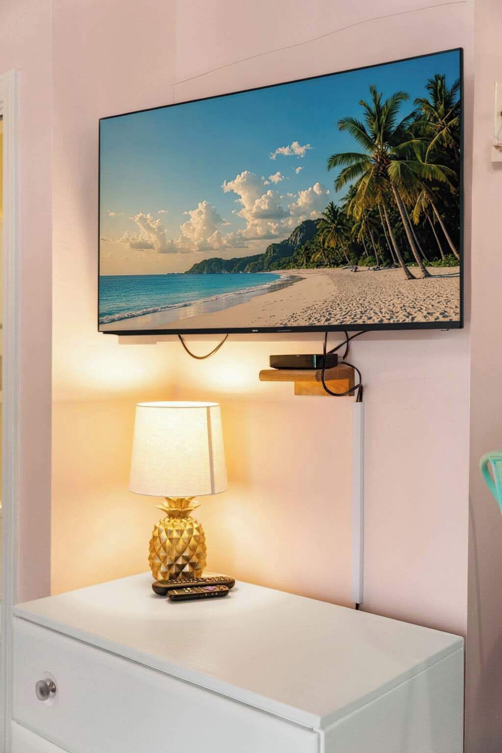 A television mounted on a wall displaying a beach scene, a white nightstand with a lamp, and a remote control.