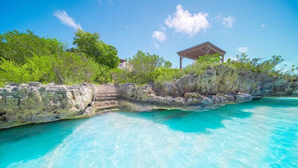 A clear turquoise pool with a rocky edge, a wooden gazebo, and lush greenery.