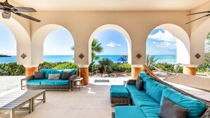 A covered outdoor seating area with wicker furniture and cushions, overlooking a beach and ocean view.