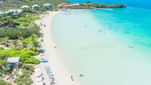 A beach with clear turquoise waters, a white sandy shore, and a few people swimming. There are several beach umbrellas and lounge chairs along the side.