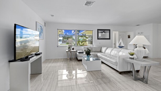 A modern living room with a flat-screen TV, a white sofa, a coffee table, and a large window offering a view of the outdoors. - Ship To Shore-Close to Beach (North Palm Beach)