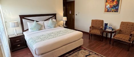 A hotel room with a large bed, two bedside tables, a nightstand, a chair, a small table with water bottles, and a painting on the wall.