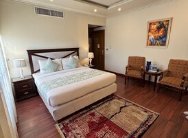 A hotel room with a large bed, two bedside tables, a nightstand, a chair, a small table with water bottles, and a painting on the wall.
