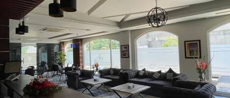 A modern hotel lobby with a reception desk, seating area with sofas and tables, large windows, and a chandelier.