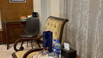 Habitación de hotel con zona de comedor que incluye una mesa de madera, una silla de cuero negro, una silla acolchada, un atril para el menú, una botella de agua y una caja de pañuelos.