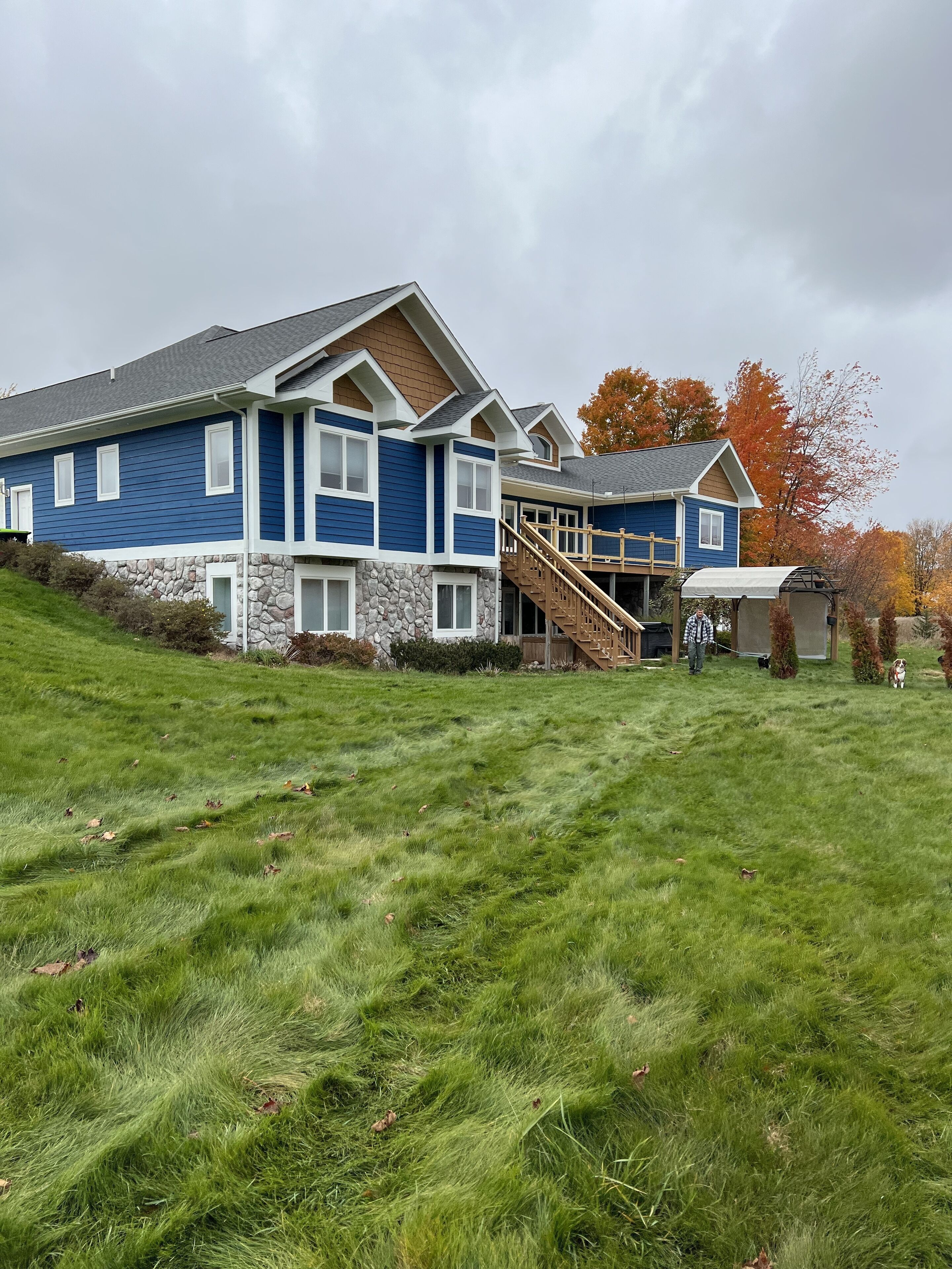 A large two-story house with a stone foundation and blue siding, a wooden deck, and a staircase leading to a covered porch.