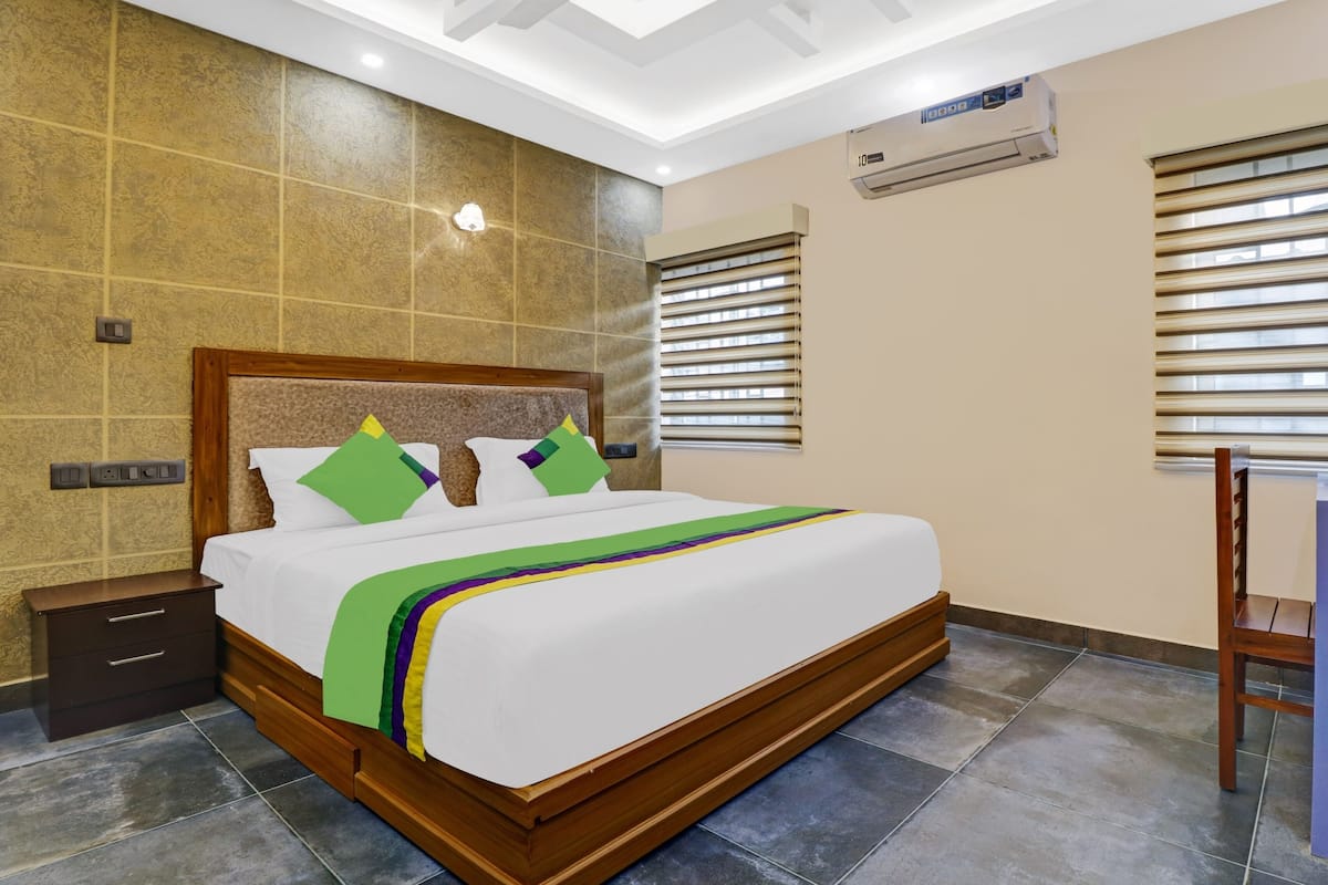Habitación de hotel con una cama grande, cabecero de madera y una mesita de noche. Se aprecia una ventana con persianas, un equipo de aire acondicionado y un escritorio con silla.
