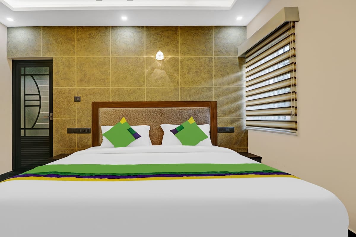 Habitación de hotel con una cama grande, cabecera, ventana con persianas y puerta con panel de vidrio.