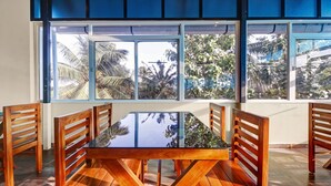 A dining area with a glass table and wooden chairs, large windows with a view of palm trees and foliage. - Treebo Pushpak 500 mtrs from Chottanikkara temple (Kanayannur)