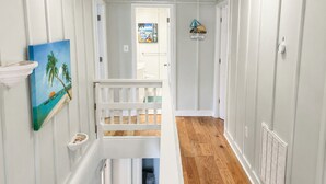 Un couloir doté d’un plancher en bois, de murs blancs, d’une rambarde blanche et d’un tableau représentant une scène de plage.