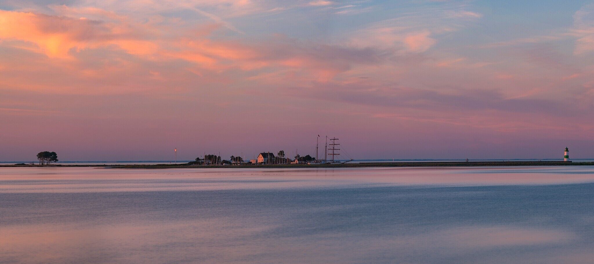 A coastal landscape with a lighthouse, calm water, and a row of buildings along the shore.
