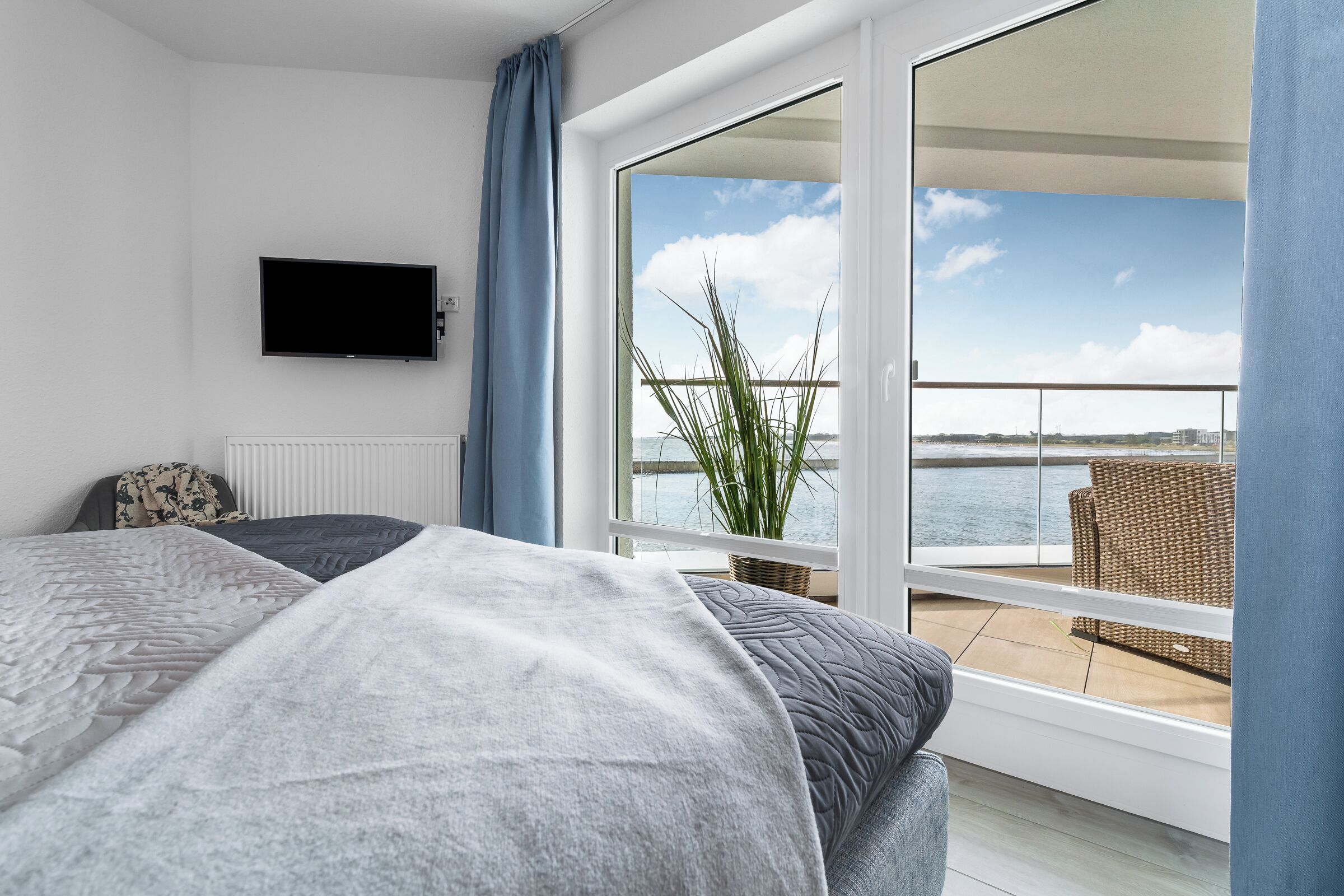A bedroom with a bed, a television, a view of the sea, and a balcony with a wicker chair.