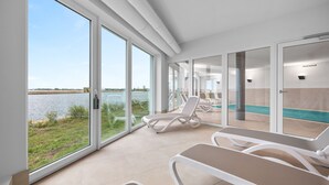 Ett modernt inomhusområde med stora fönster som vetter mot en vattenyta, med solstolar och en pool.