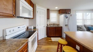 Una cocina funcional con microondas, estufa, pileta y refrigerador. Cuenta con alacenas de madera, una mesada de granito y una pequeña isla con taburetes.