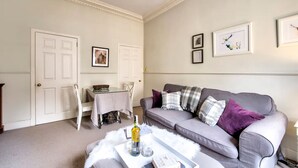 A living room with a grey sofa, a white door, a dining table with chairs, and framed pictures on the wall. - JOIVY 1-Bed Flat In Edinburgh Old Town (Edinburgh)