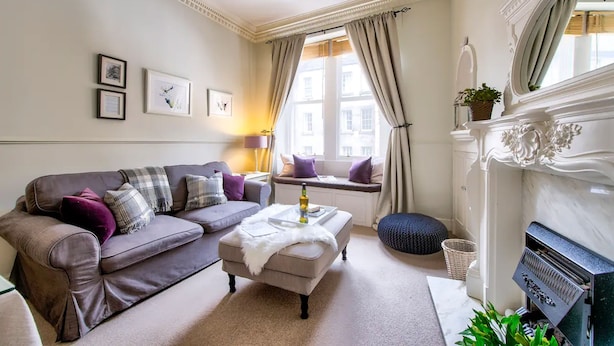 A living room with a sofa, ottoman, and a fireplace. - JOIVY 1-Bed Flat In Edinburgh Old Town (Edinburgh)