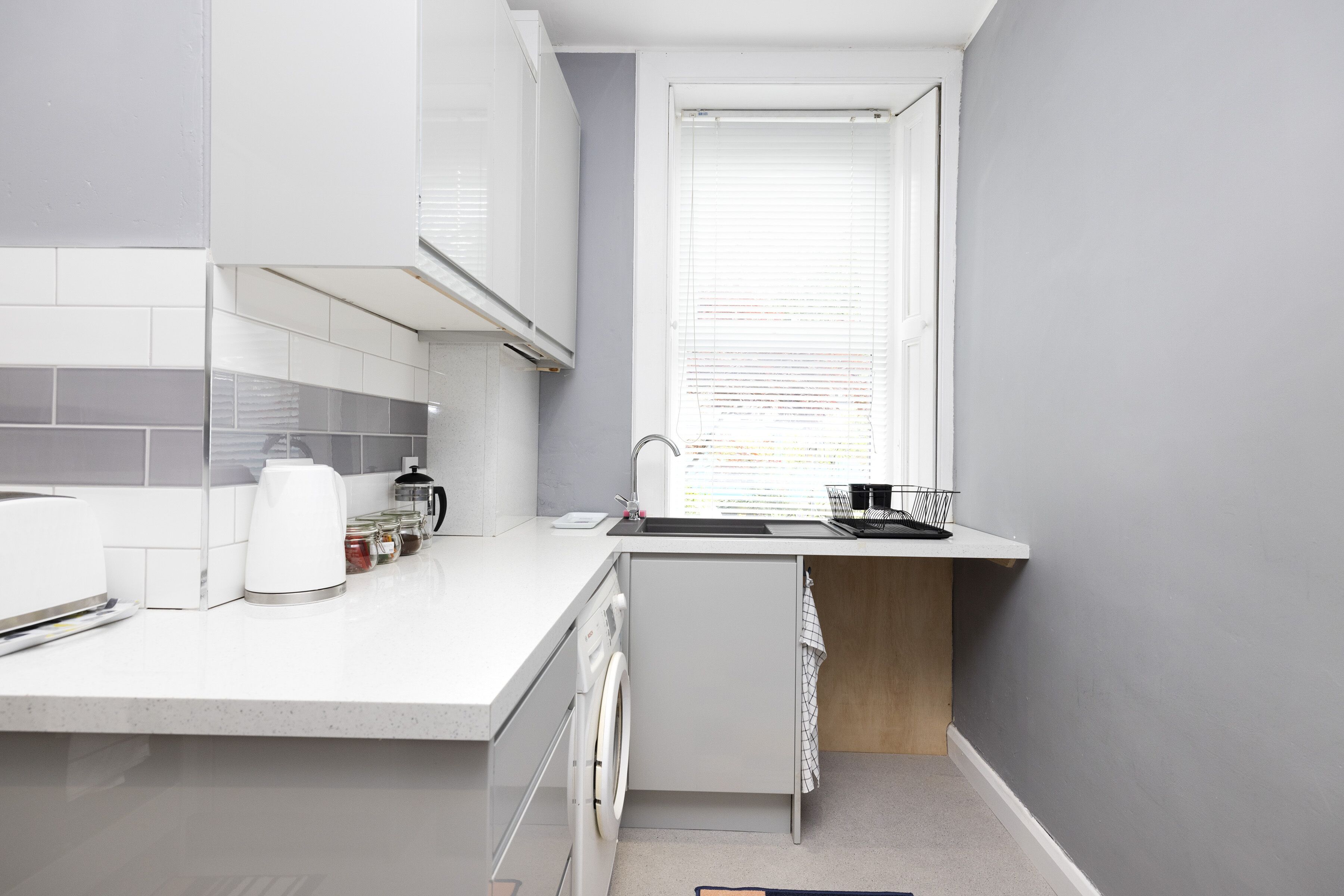 A modern kitchen with a built-in washing machine, a coffee maker, and a window with blinds.
