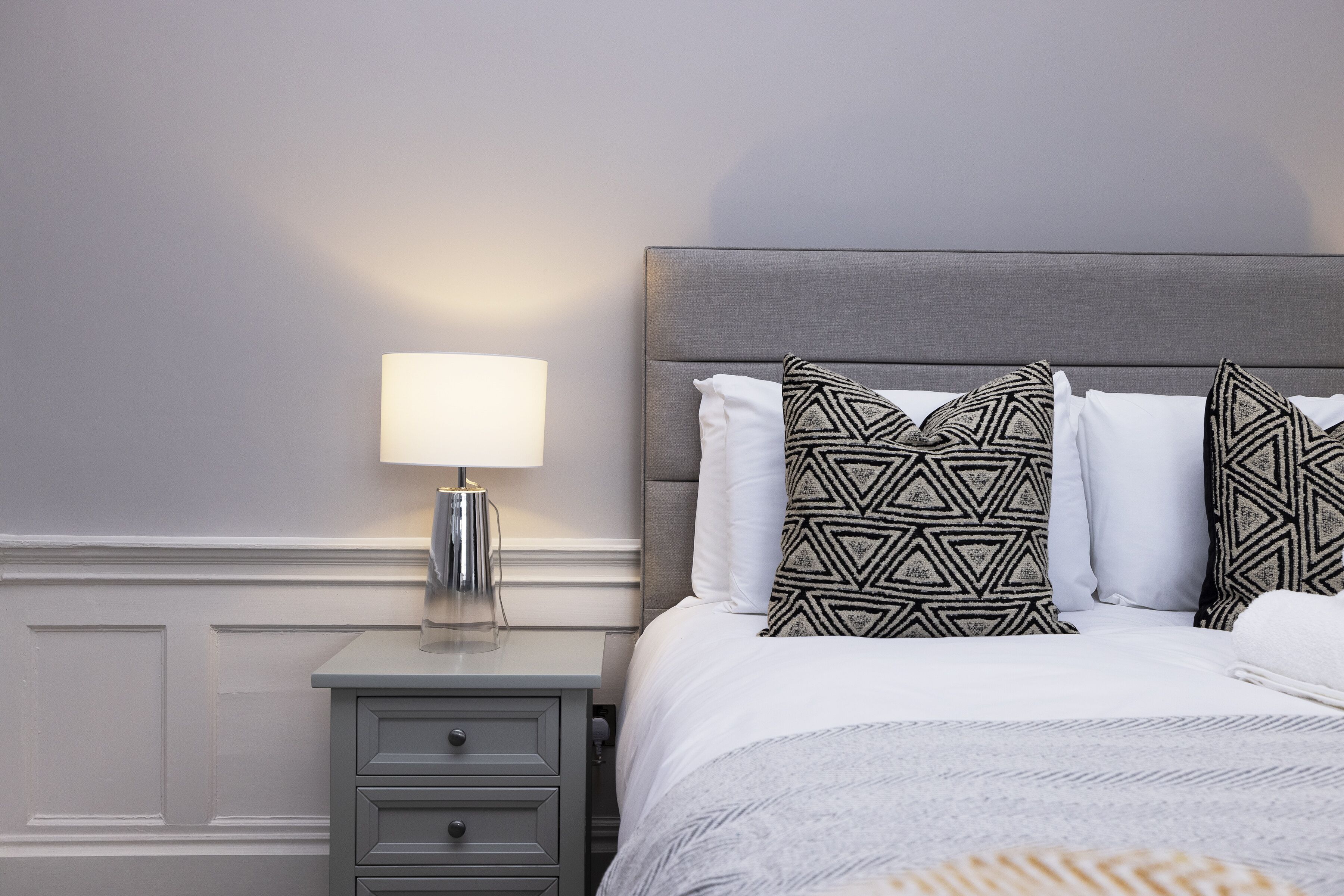 Un lit bien fait, agrémenté d’oreillers à motifs, une lampe de chevet et une petite commode.