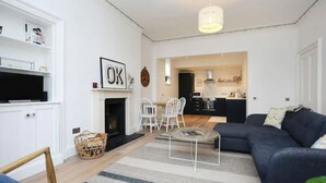 A modern living room with a dark sofa, a glass coffee table, and a fireplace. There is a kitchen visible through an open doorway. - JOIVY Luxury House With Patio Next To North Berwick Beach (North Berwick)