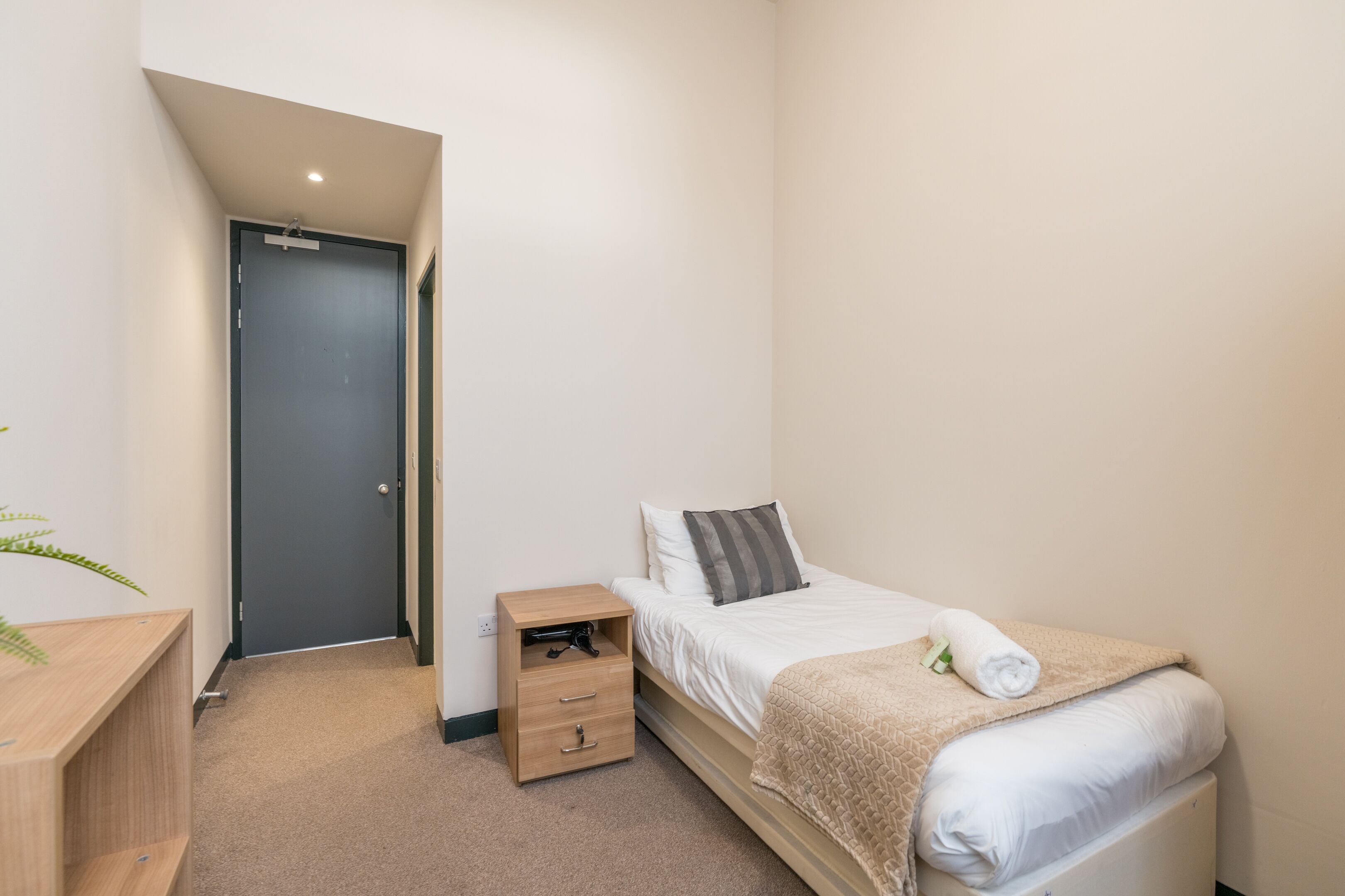Un dormitorio pequeño y ordenado con una cama individual, una mesita de noche y una puerta que da a otra habitación.