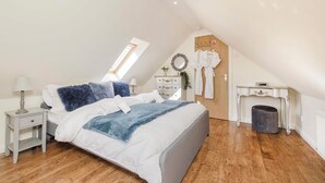 A bedroom with a sloped ceiling, a bed with white and blue bedding, a nightstand with a lamp, a dresser, and a small table with a chair. - JOIVY Greenknowes Cottage W/ Hot Tub & Fireplace (Kelty)
