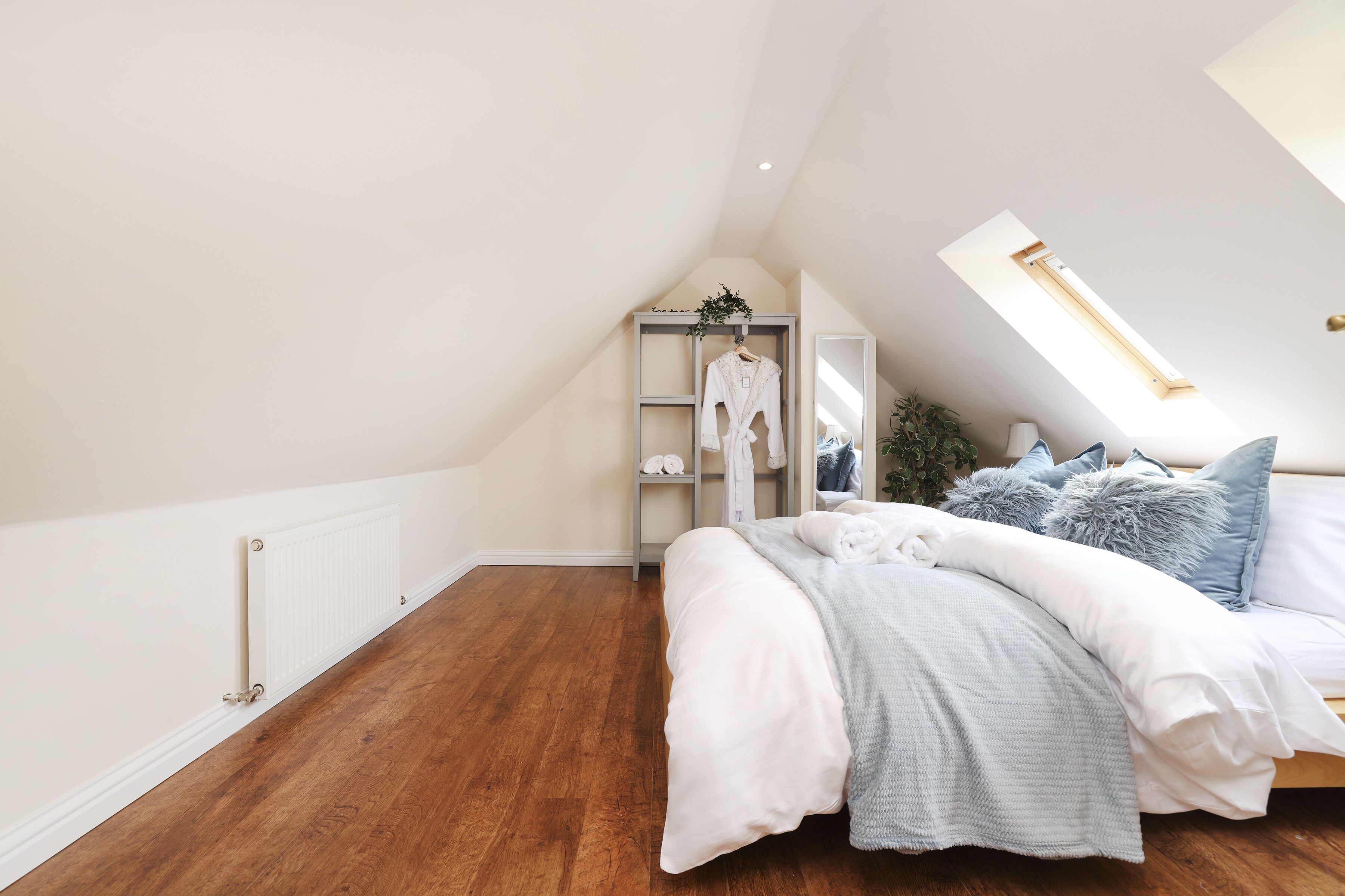 A bedroom with a sloped ceiling, a skylight, a bed with white bedding, a grey blanket, and a grey wardrobe with clothes hanging inside.