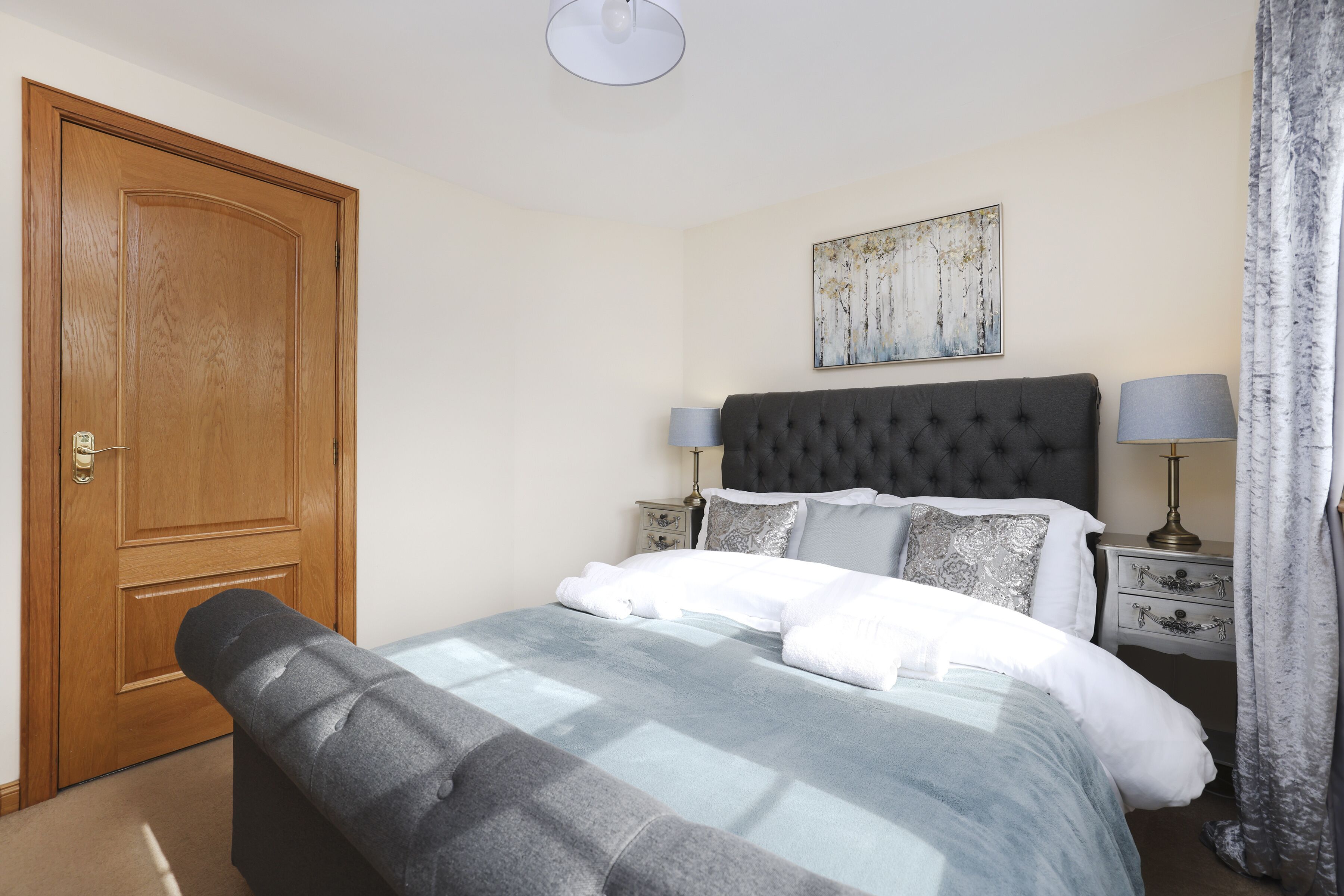 A neatly arranged bedroom with a large bed, a wooden door, a bedside table with lamps, and a wall-mounted artwork.